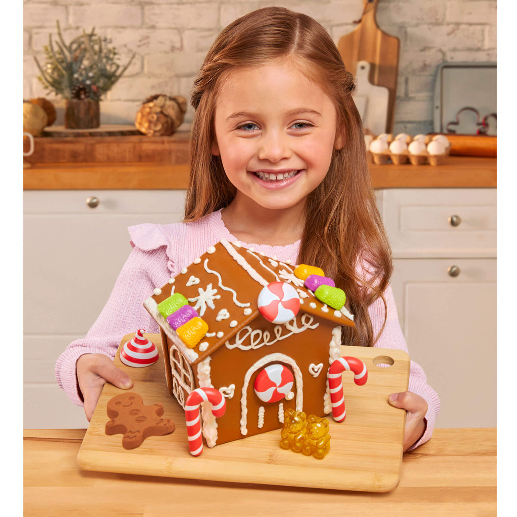 Young girl holding a gingerbread house she made in a kitchen.